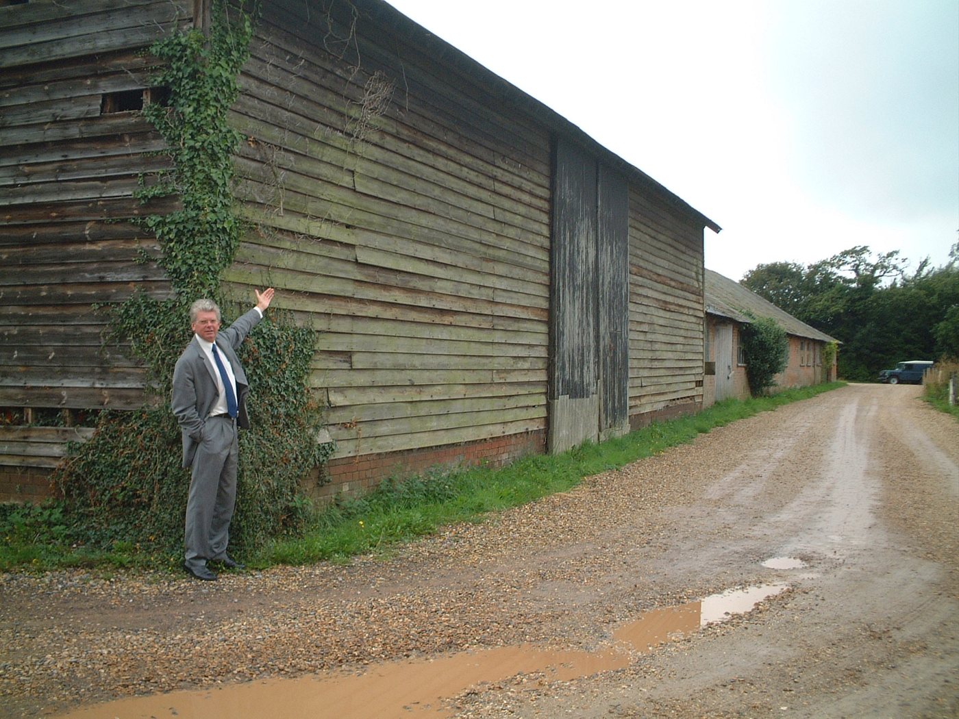 A man in a grey suit raises an arm towards a large, ivy-covered weatherboarded barn beside a gravel track