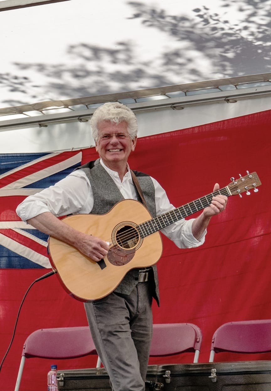 David Morgan Walters, white-haired and smiling, playing an acoustic guitar on a stage in front of a red flag