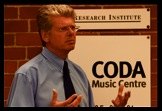 Dave Walters in a blue shirt and tie, speaking in front of a sign for the Research Institute and Coda Music Centre