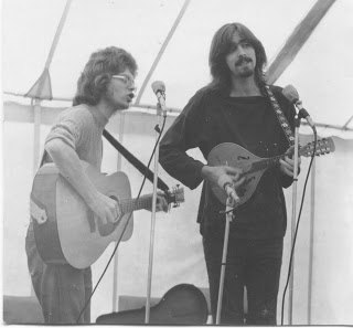Black and white photograph of two musicians on a stage in a marquee: one plays an acoustic guitar, the other a mandolin